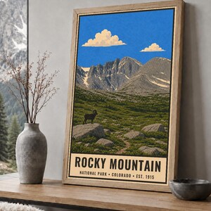 May include: A framed vertical poster featuring a landscape illustration of Rocky Mountain National Park, Colorado. The artwork depicts a bighorn sheep on a rock, green terrain, jagged mountains, and blue sky with clouds. Text reads: ROCKY MOUNTAIN NATIONAL PARK COLORADO EST. 1915.