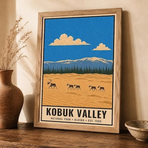 Op de afbeelding: Een ingelijste kunstdruk met een landschap van Kobuk Valley National Park in Alaska. De illustratie toont een helderblauwe lucht met wolken, besneeuwde bergen, een donker bos en een kudde kariboes op een zandkleurig veld met tekst.