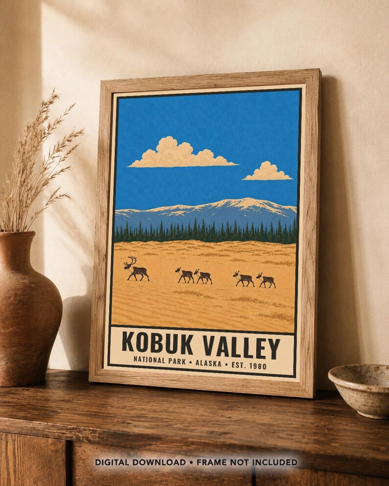 Op de afbeelding: Een ingelijste kunstdruk met een landschap van Kobuk Valley National Park in Alaska. De illustratie toont een helderblauwe lucht met wolken, besneeuwde bergen, een donker bos en een kudde kariboes op een zandkleurig veld met tekst.