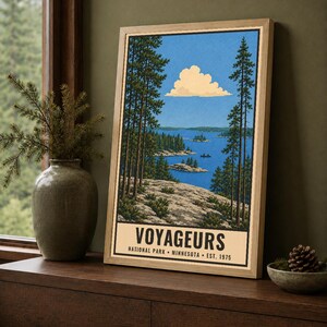 Op de afbeelding: Een ingelijste verticale kunstdruk met een landschapsillustratie van Voyageurs National Park in Minnesota. De afbeelding toont een rotsachtige kustlijn, hoge dennenbomen, een blauw meer met kleine eilanden en een wolk. De tekst luidt VOYAGEURS NATIONAL PARK MINNESOTA EST. 1975.
