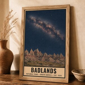 K&ouml;nnte beinhalten: Ein gerahmter vertikaler Wandkunstdruck des Badlands Nationalparks in South Dakota. Das Bild zeigt einen Sternenhimmel mit der Milchstra&szlig;e &uuml;ber zerkl&uuml;fteten, geschichteten Felsformationen. Der Text lautet BADLANDS NATIONAL PARK SOUTH DAKOTA EST. 1978.