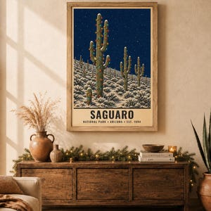 May include: A rectangular wall art print featuring a dark blue night sky over a desert landscape with snow-covered ground and saguaro cacti decorated with colorful string lights. The text reads SAGUARO NATIONAL PARK ARIZONA EST. 1994.