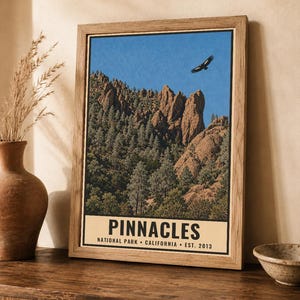 Op de afbeelding: Een ingelijste wandkunstprint met een landschap van Pinnacles National Park in Californi&euml;. De afbeelding toont rotsachtige kliffen, dennenbomen en een vogel die in een blauwe lucht vliegt. De tekst luidt PINNACLES NATIONAL PARK CALIFORNIA EST. 2013.