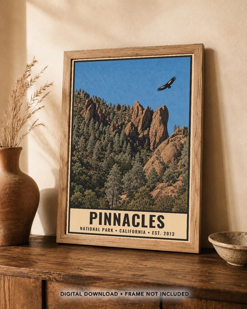 Op de afbeelding: Een ingelijste wandkunstprint met een landschap van Pinnacles National Park in Californi&euml;. De afbeelding toont rotsachtige kliffen, dennenbomen en een vogel die in een blauwe lucht vliegt. De tekst luidt PINNACLES NATIONAL PARK CALIFORNIA EST. 2013.