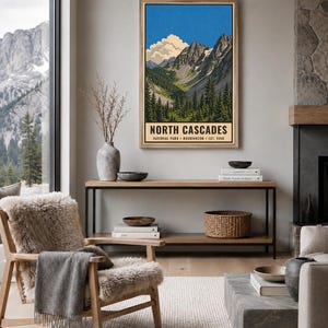 Op de afbeelding: Een ingelijste verticale wandkunstprint van het North Cascades National Park in Washington, opgericht in 1968. De illustratie toont grillige bergtoppen, een helderblauwe lucht met witte wolken en een dicht bos met groene dennenbomen.