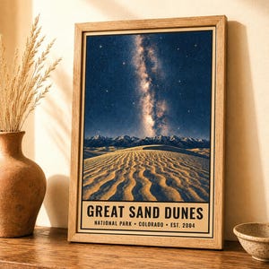 May include: A framed vertical poster featuring a night sky with a visible Milky Way galaxy above a desert landscape of sand dunes and distant mountains. The text at the bottom reads GREAT SAND DUNES NATIONAL PARK COLORADO EST. 2004.