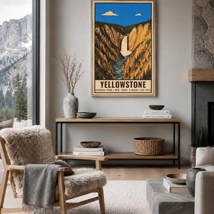 Op de afbeelding: Een ingelijste verticale wandkunstprint met een gestileerde illustratie van Yellowstone National Park met een waterval door een kloof, blauwe lucht met wolken en de tekst Yellowstone National Park, Wyo., Mont. & Idaho Est. 1872.
