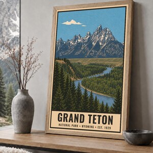 K&ouml;nnte beinhalten: Ein gerahmtes vertikales Poster mit einer Landschaftsillustration des Grand-Teton-Gebirges mit schneebedeckten Gipfeln, einem gewundenen blauen Fluss und dichten gr&uuml;nen Kiefernw&auml;ldern. Der Text lautet GRAND TETON NATIONAL PARK WYOMING EST. 1929.