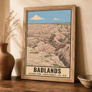 May include: A framed vertical art print featuring a landscape illustration of Badlands National Park in South Dakota. The image displays layered rock formations in shades of tan, pink, and brown under a blue sky with clouds. Text reads BADLANDS.