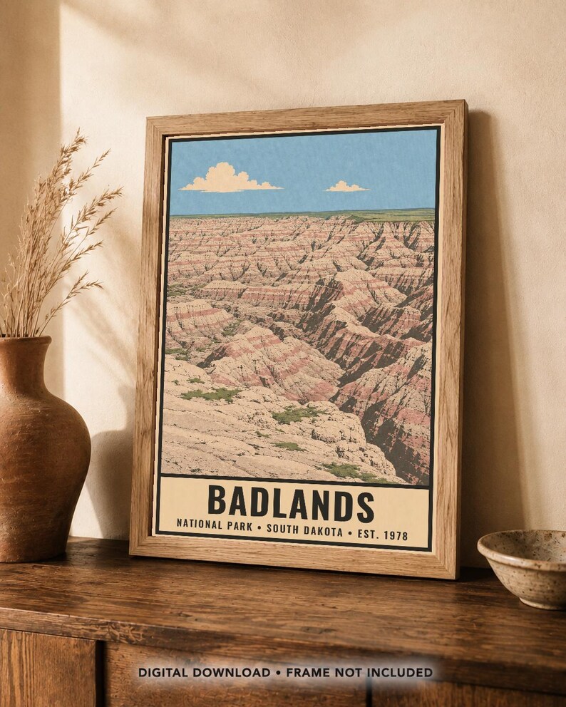 May include: A framed vertical art print featuring a landscape illustration of Badlands National Park in South Dakota. The image displays layered rock formations in shades of tan, pink, and brown under a blue sky with clouds. Text reads BADLANDS.