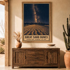 May include: A vertical wall poster featuring a night sky with the Milky Way galaxy above the Great Sand Dunes, with text reading GREAT SAND DUNES NATIONAL PARK COLORADO EST. 2004. The poster is displayed in a wooden frame on a wall.