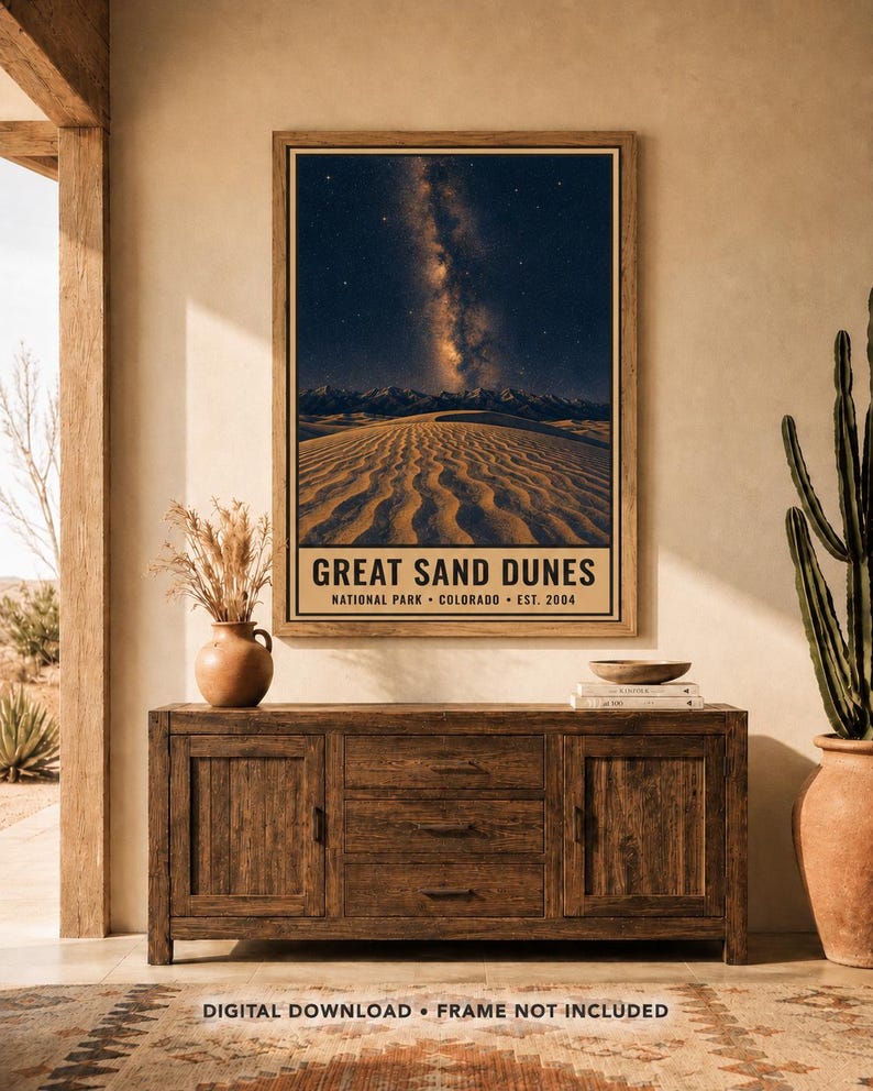 May include: A vertical wall poster featuring a night sky with the Milky Way galaxy above the Great Sand Dunes, with text reading GREAT SAND DUNES NATIONAL PARK COLORADO EST. 2004. The poster is displayed in a wooden frame on a wall.
