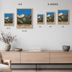 Op de afbeelding: Een collectie van vijf ingelijste kunstprints met het North Cascades National Park met berg- en boslandschappen. De prints zijn gelabeld met de maten 8x10, 16x20, A4, A3 en A2. De tekst luidt North Cascades National Park Washington Est 1968.