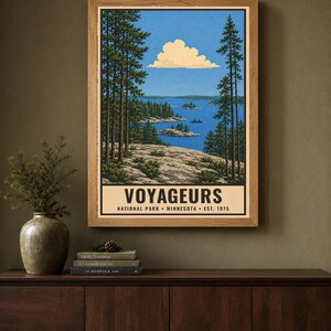 Op de afbeelding: Een ingelijste verticale poster met een landschapsillustratie van Voyageurs National Park in Minnesota. Het kunstwerk toont een blauw meer, een rotsachtige kustlijn, hoge dennenbomen en een enkele wolk. De tekst luidt VOYAGEURS NATIONAL PARK MINNESOTA EST. 1975.