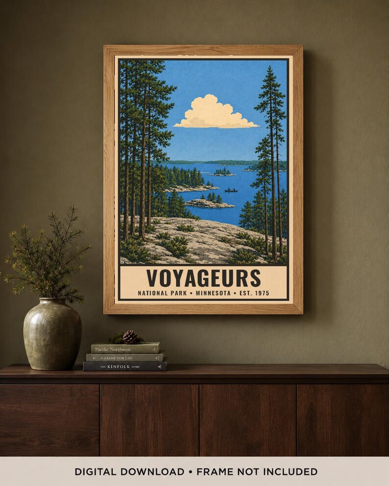 Op de afbeelding: Een ingelijste verticale poster met een landschapsillustratie van Voyageurs National Park in Minnesota. Het kunstwerk toont een blauw meer, een rotsachtige kustlijn, hoge dennenbomen en een enkele wolk. De tekst luidt VOYAGEURS NATIONAL PARK MINNESOTA EST. 1975.