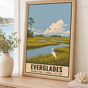 K&ouml;nnte beinhalten: Ein gerahmtes vertikales Poster mit einer Landschaftsillustration des Everglades-Nationalparks in Florida. Das Kunstwerk zeigt einen wei&szlig;en Reiher in einem grasbewachsenen Sumpfgebiet mit einem gewundenen blauen Wasserlauf, hohen B&auml;umen und Wolken.
