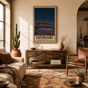 May include: A vertical wall art print featuring a night sky over the rock arch formation in Canyonlands National Park, Utah. The text reads CANYONLANDS NATIONAL PARK UTAH EST. 1964. The print is displayed in a wooden frame on a beige wall.
