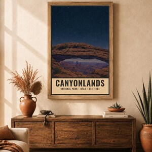 May include: A vertical rectangular wall art print featuring a night sky over a natural rock arch in Canyonlands National Park, Utah. The text reads CANYONLANDS NATIONAL PARK UTAH EST. 1964. The print is displayed in a wooden frame on a wall.