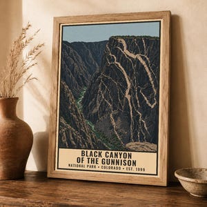 Op de afbeelding: Een ingelijste poster met een grafische illustratie van de Black Canyon of the Gunnison met een kronkelende rivier aan de basis en steile kliffen. De tekst luidt: BLACK CANYON OF THE GUNNISON NATIONAL PARK COLORADO EST. 1999. De lijst is van licht hout.
