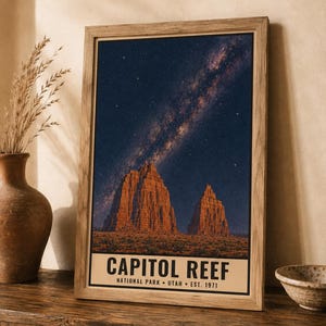 May include: A framed vertical poster featuring a night sky filled with stars and the Milky Way galaxy above two large, orange-toned rock formations. The bottom of the print displays the text CAPITOL REEF NATIONAL PARK UTAH EST. 1971 in black.