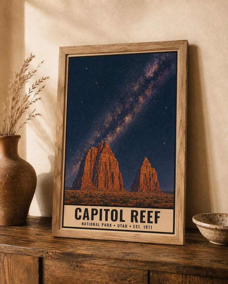 May include: A framed vertical poster featuring a night sky filled with stars and the Milky Way galaxy above two large, orange-toned rock formations. The bottom of the print displays the text CAPITOL REEF NATIONAL PARK UTAH EST. 1971 in black.