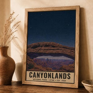 May include: A framed vertical poster featuring a night sky filled with stars above a natural rock arch and canyon landscape. The bottom section contains the text CANYONLANDS NATIONAL PARK UTAH EST. 1964 in black capital letters on a beige background.