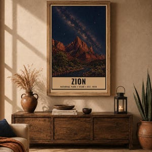 May include: A rectangular wall art print featuring a night landscape of Zion National Park with red rock mountains under a starry sky and the text ZION NATIONAL PARK UTAH EST. 1919. The print is displayed in a wooden frame on a beige wall.