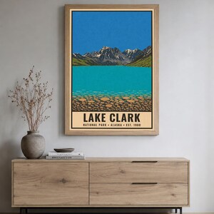 Pu&ograve; includere: Un poster verticale incorniciato raffigurante un'illustrazione paesaggistica del Lake Clark National Park in Alaska. L'opera mostra montagne innevate, un cielo azzurro brillante, un lago turchese e un primo piano roccioso con il testo LAKE CLARK.