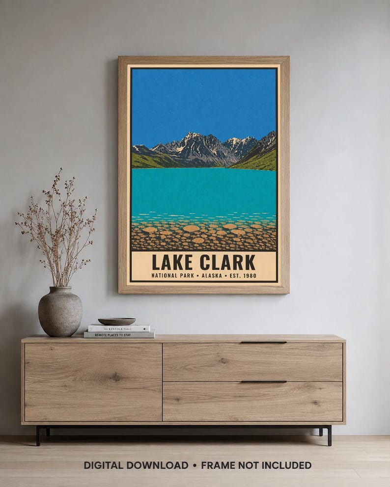 Pu&ograve; includere: Un poster verticale incorniciato raffigurante un'illustrazione paesaggistica del Lake Clark National Park in Alaska. L'opera mostra montagne innevate, un cielo azzurro brillante, un lago turchese e un primo piano roccioso con il testo LAKE CLARK.