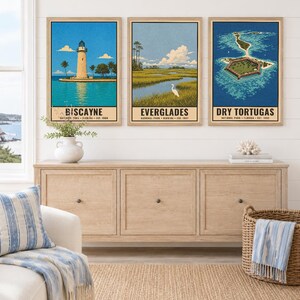 May include: Three framed vertical wall art prints featuring illustrations of Florida national parks. The left print shows the Biscayne lighthouse, the center print depicts the Everglades wetlands with a white bird, and the right print shows Dry Tortugas.