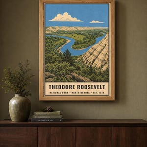 May include: A rectangular wall art print in a wooden frame depicting a landscape of Theodore Roosevelt National Park in North Dakota. The illustration features a winding blue river, green forests, and layered rock formations under a blue sky.