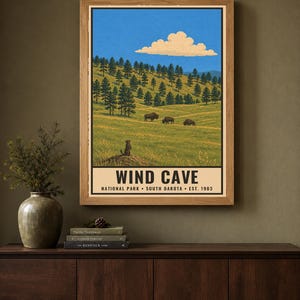 May include: A framed poster featuring a landscape illustration of Wind Cave National Park in South Dakota. The artwork depicts a grassy hillside with pine trees, three bison, and a prairie dog, under a blue sky with a single cloud. Text reads WIND CAVE.