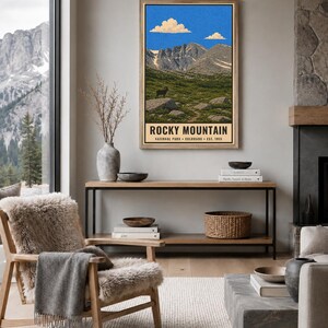 May include: A vertical wall art print featuring a landscape illustration of Rocky Mountain National Park in Colorado. The artwork depicts jagged mountain peaks under a blue sky with clouds, a green meadow, and a bighorn sheep. Text reads ROCKY MOUNTAIN NATIONAL PARK COLORADO EST. 1915.