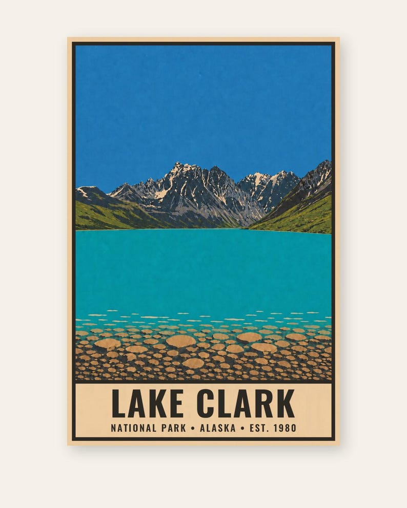 Pu&ograve; includere: Un poster rettangolare con un'illustrazione paesaggistica del Lake Clark in Alaska. La scena mostra un cielo azzurro brillante, montagne frastagliate innevate, colline verdi e un lago turchese con pietre rotonde visibili sul fondo. Il testo recita LAKE CLARK NATIONAL PARK ALASKA EST. 1980.