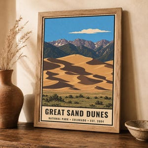 K&ouml;nnte beinhalten: Ein gerahmtes Poster mit einer stilisierten Illustration des Great Sand Dunes Nationalparks in Colorado. Das Kunstwerk zeigt goldene Sandd&uuml;nen, gr&uuml;ne Vegetation und schneebedeckte Berge unter einem blauen Himmel mit Text am unteren Rand.