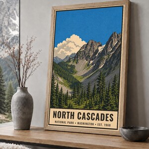 Op de afbeelding: Een ingelijste verticale poster met een gestileerde illustratie van het North Cascades National Park in Washington. Het kunstwerk toont grillige bergtoppen, een dicht naaldbos en een grote witte wolk tegen een helderblauwe lucht.