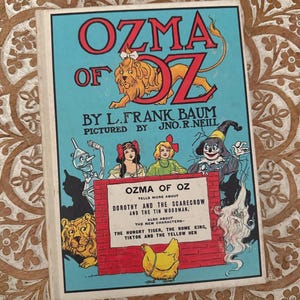 May include: A hardcover book titled Ozma of Oz by L. Frank Baum, illustrated by Jno. R. Neill. The light blue cover features a lion, the Tin Woodman, Dorothy, the Scarecrow, and a yellow hen, with a red brick wall graphic containing text.