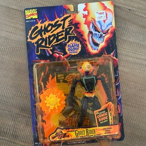 Może przedstawiać: Figurkę akcji Ghost Ridera z elementami świecącymi w ciemności. Figurka jest w pozie z płonącym łańcuchem, a w zestawie znajduje się komiks. Opakowanie jest czarne i pomarańczowe z logo Ghost Ridera.