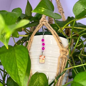 May include: A decorative hanging ornament featuring three bright pink spherical beads and a small rectangular charm. The charm displays a light-colored illustration of a potted plant with the text "I'm plant happy" printed in a dark brown font.