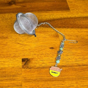 May include: A stainless steel mesh tea infuser ball attached to a silver-toned chain. The chain features alternating green beads and silver flower-shaped spacers, ending in a yellow and pink enamel charm shaped like a honey pot with the word HONEY.