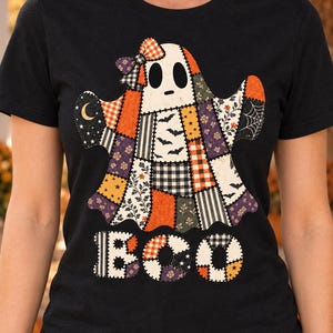 May include: A black short-sleeve t-shirt featuring a large ghost graphic made of various patchwork patterns including orange, black, and white plaid, stars, bats, and floral prints. A bow sits on the ghost's head, and the word BOO is printed below.