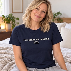 May include: A navy blue short-sleeved t-shirt featuring the white text "I'd rather be napping" centered on the chest, accompanied by a simple white line-art graphic of a bed positioned directly underneath the printed text.