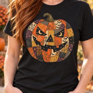 Op de afbeelding: Een zwart T-shirt met korte mouwen en een grote afbeelding van een Halloween-pompoen. Het pompoenontwerp is samengesteld uit verschillende patchwork-stofpatronen in oranje, zwart en geel, inclusief motieven van sterren, vleermuizen en spinnenwebben.