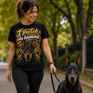 May include: A black short-sleeve t-shirt featuring a graphic of a Doberman dog, paw prints, a heart, and the text I protek dis human in yellow and white lettering. The design includes golden light streaks radiating from the dog illustration.
