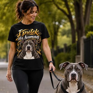May include: A black short-sleeve t-shirt featuring a graphic of a gray and white pit bull dog with gold paw prints and the text I protek dis human in gold and white lettering. The shirt is paired with black leggings and a black dog leash.