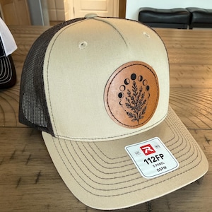 Peut inclure: Une casquette trucker en maille beige et marron foncé avec un écusson en cuir circulaire orné d'un motif botanique et de phases lunaires noires. Une étiquette blanche sur la visière porte le texte R 112FP 5 PANEL OSFM. Panneau avant structuré.