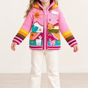May include: A pink knit zip-up hoodie featuring a colorful landscape scene with embroidered animals, a sun, and a cloud. The sleeves have horizontal stripes in yellow, orange, green, and purple. The front includes two pockets and a full-length zipper.