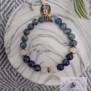 Peut inclure: Un bracelet composé de perles rondes en pierre bleue et blanche marbrée, alternées avec des perles blanches translucides et des intercalaires en métal doré. Un petit pendentif rond à bordure dorée avec une illustration pend au sommet.