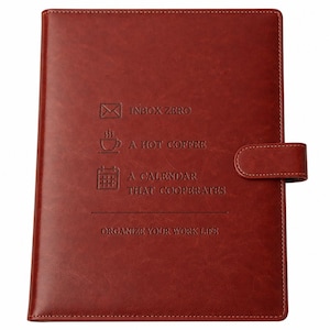 May include: A dark red leather-like notebook with a magnetic strap closure. The cover features embossed icons and text: INBOX ZERO, A HOT COFFEE, A CALENDAR THAT COOPERATES, and ORGANIZE YOUR WORK LIFE. The surface has a smooth, consistent texture.