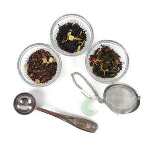 May include: Three small glass bowls filled with loose leaf tea blends, a silver tea infuser, a silver measuring spoon with the text "Perfect Cup of Tea" and a jade green crescent moon charm.